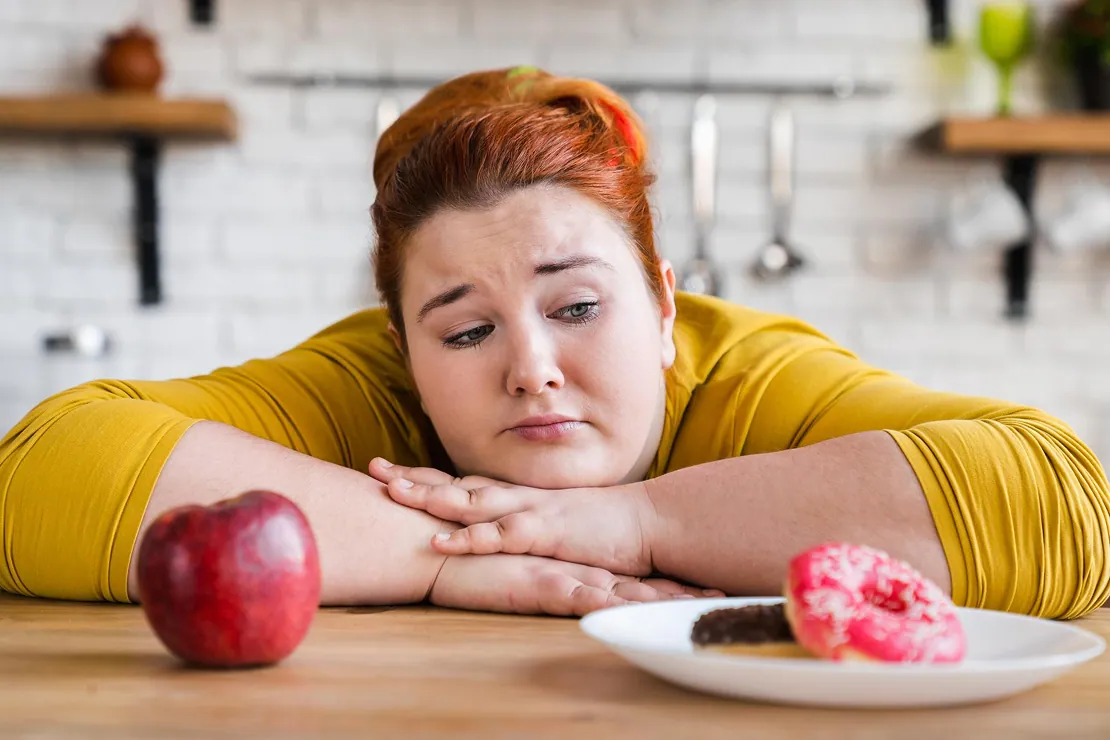 A person looking sad while contemplating an apple and a donut, symbolizing the mental struggle in weight loss.